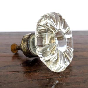 Può includere: Un pomello per mobile in vetro trasparente caratterizzato da un design circolare, scanalato e floreale con un centro liscio. Il pomello è fissato a una base metallica scura e ossidata con una vite filettata che sporge dal retro.