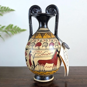 May include: A hand-painted ceramic vase with a black rim and handles. The vase features a cream-colored background with red and black illustrations of birds, a horse, and figures. A tag hangs from the vase.