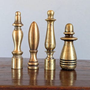 Pode incluir: Quatro terminais de l&acirc;mpada antigos de lat&atilde;o em v&aacute;rias formas, incluindo uma urna cl&aacute;ssica, uma bala e um design escalonado. Os terminais s&atilde;o exibidos em uma superf&iacute;cie de madeira escura, mostrando sua p&aacute;tina envelhecida e detalhes intrincados.