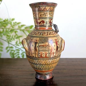 May include: A small, antique-style vase with a detailed, hand-painted design. The vase features a cream-colored base with red, brown, and black accents. It has a bulbous body, two handles, and a narrow neck, showcasing ancient Greek-inspired artwork.