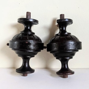May include: Two dark brown, antique-style finials. Each finial has a rounded, bulbous center section, a slender neck, and a flared base. A short, cylindrical post extends from the top of each finial. The finials are likely made of wood.