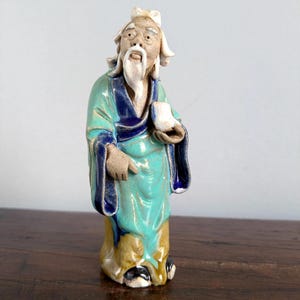 Puede incluir: Figura de cerámica de un anciano con vestimenta asiática tradicional. La figura lleva una túnica turquesa con ribetes azul oscuro, una larga barba blanca y sostiene una pequeña taza blanca. La base es amarilla y la figura tiene zapatos negros.