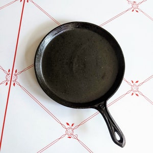 May include: A black cast iron griddle pan with a single handle. The pan is round with a flat cooking surface. The handle has a hole for hanging. The pan is on a white surface with a red floral pattern.