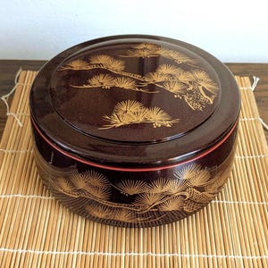 May include: A circular, dark brown lacquered box with a lid, decorated with a gold-coloured pine tree design. The box has a red accent around the rim and sits on a bamboo mat. The lid and sides are decorated with the pine tree motif.