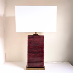 May include: A rectangular table lamp with a red textured base and a white rectangular lampshade. The base has a stacked, ribbed design. The lamp sits on a square, gold-colored base. The lamp is a decorative lighting fixture.