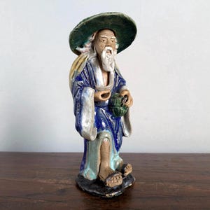 Puede incluir: Figura de cerámica de un anciano con túnica azul y sombrero verde ancho. La figura tiene una larga barba blanca y sostiene un recipiente verde. La figura está sobre una base negra.