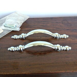May include: Two polished chrome drawer pulls featuring a curved arch design with decorative finial ends. Each handle has a smooth, reflective metallic finish and includes two small metal mounting screws enclosed in a clear plastic bag.