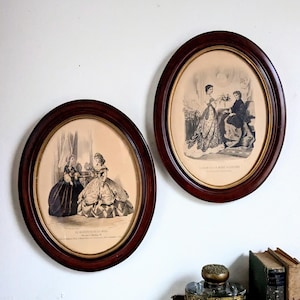 Könnte beinhalten: Zwei ovale Rahmen aus dunklem Holz mit schwarz-weißen Modeillustrationen. Der linke Druck trägt den Titel LE MONITEUR DE LA MODE und der rechte den Titel ALBUM DE LA MODE ILLUSTREE, beide zeigen Figuren in historischer Kleidung.
