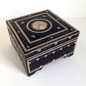 May include: A rectangular black box featuring intricate gold-colored inlay patterns. The lid displays a central circular medallion with a detailed relief design, surrounded by decorative borders, floral motifs, and scrolling line work.