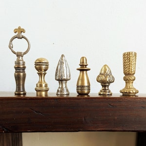 May include: Six decorative finials in brass and silver tones, displayed on a dark wooden surface. The finials have different shapes, suitable for lamps or furniture. The background is white. The finials are made of metal.