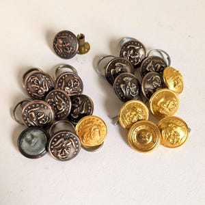 May include: A collection of vintage metal buttons in various colors. The buttons feature detailed embossed designs, including eagles and classical figures. Some buttons are bronze, others are gold, and some have a silver-grey finish. A small brass tube is also present.
