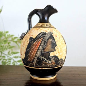 Peut inclure: Une cruche en céramique avec une anse et une base noires sur un corps de couleur crème. Le récipient présente une illustration de profil en style figures noires représentant une personne coiffée, ornée d'un motif grec géométrique au col.