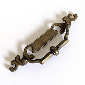 May include: A brass-toned metal drawer pull featuring an elongated backplate with decorative scrollwork at both ends and a central rectangular section. A hinged, faceted drop handle is attached to the backplate, allowing for movement.