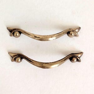 Op de afbeelding: Twee zilverkleurige metalen ladehandgrepen met een gebogen, boogvormig ontwerp. Elke handgreep heeft een gladde, centrale stang die taps toeloopt naar de uiteinden en eindigt in een klein, afgerond bolvormig detail bij elk bevestigingspunt, afgewerkt met een metaalglans.