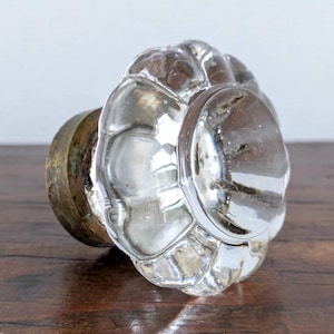 May include: A clear glass cabinet knob featuring a multi-faceted, flower-like scalloped edge design. The knob is attached to a circular, tarnished brass-toned metal base that connects to the back, providing a contrast to the transparent glass.
