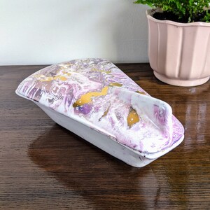 May include: A decorative ceramic trinket box with a lid. The lid has a marbled design in shades of purple, white, and gold. The base of the box is light grey. The box is on a dark wood surface.