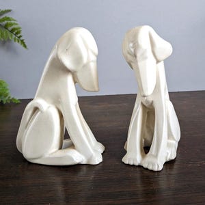 May include: Two off-white ceramic dog figurines. The dogs are in a seated position with their heads bowed. The figurines have a smooth, glossy finish and are placed on a dark wooden surface. The background is a muted gray.