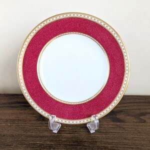 May include: A round porcelain plate featuring a plain white center surrounded by a wide, textured magenta border. The rim is decorated with a gold-colored band and a repeating white and gold geometric pattern along the outer edge of the plate.