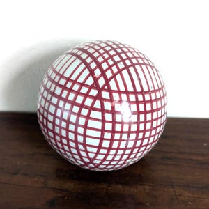 Puede incluir: Un objeto esférico con una superficie blanca lisa y brillante que presenta un patrón de cuadrícula repetitivo de líneas de color rojo oscuro que se cruzan y envuelven toda la circunferencia de la bola, creando un diseño geométrico uniforme.
