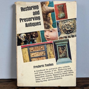 May include: Vintage book titled "Restoring and Preserving Antiques" by Frederic Taubes. The cover features a collage of antique objects, including a sculpture, vase, and a small chest of drawers. The book is a handbook for art restoration.