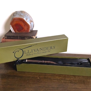 May include: A brown wooden wand rests inside a rectangular olive green box with a black interior lining. The lid features the black text Ollivanders Makers of Fine Wands since 382 BC with a stylized wand graphic. The box is open and displayed.