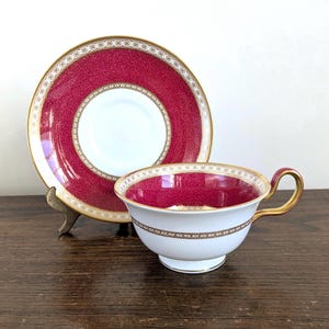 May include: A white porcelain teacup and saucer set featuring a vibrant magenta pink textured pattern. The rim of the saucer and the interior of the cup are decorated with this pink design, accented by intricate gold filigree borders and a gold handle.