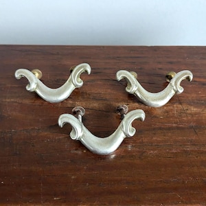 May include: Three silver-toned drawer pulls with a curved, ornate design. Each pull has a brass-colored screw head. The pulls are arranged on a dark brown wooden surface.