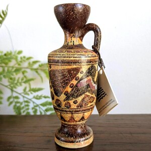 May include: A small, brown ceramic vase with a handle and a flared top. The vase features intricate patterns in shades of brown, orange, and black. A tag hangs from the handle. The vase is displayed on a wooden surface.