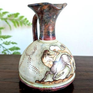 May include: A small, antique-style ceramic jug with a handle and a flared rim. The jug features a hand-painted scene of figures and a horse in shades of brown, beige, and red. The jug is displayed on a wooden surface.