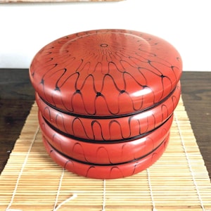 May include: A stack of three round, red lacquer containers with a glossy finish. Each container features a hand-painted black wavy line pattern that radiates from the center of the lid and continues down the sides of the stacked circular vessels.