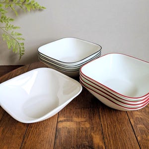 Puede incluir: Un conjunto de cuencos cuadrados blancos con bordes de colores. Algunos cuencos tienen ribetes negros, verdes y rojos. Los cuencos están apilados en juegos y colocados sobre una superficie de madera. Una planta verde es visible en el fondo.