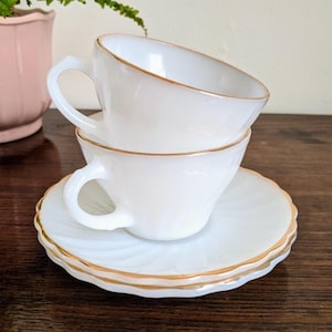 May include: Two white teacups with gold trim are stacked on a white saucer with a gold rim. The teacups have curved handles. The saucer has a swirled design. The teacups and saucer are on a dark wooden surface.