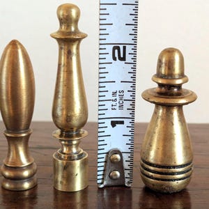 Pode incluir: Tr&ecirc;s terminais de lat&atilde;o antigos de v&aacute;rias formas e tamanhos s&atilde;o exibidos ao lado de uma r&eacute;gua. O terminal mais alto tem aproximadamente 5 cm de altura. Os terminais t&ecirc;m uma p&aacute;tina quente e envelhecida, sugerindo que s&atilde;o vintage ou antigos.