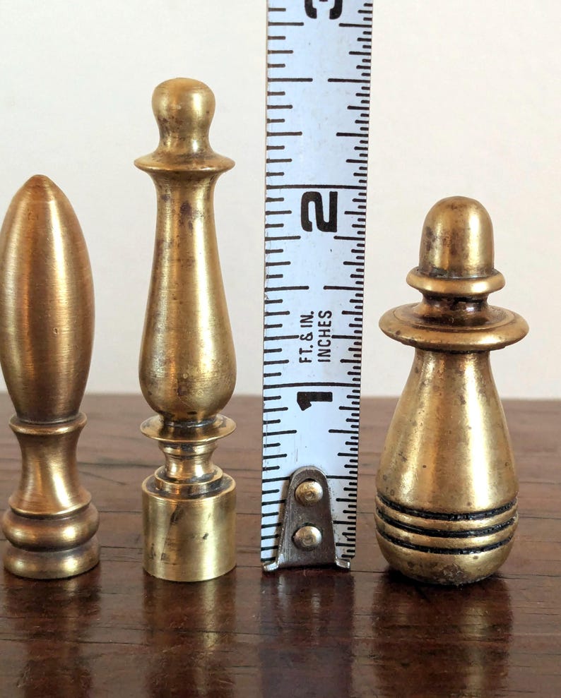 Pode incluir: Tr&ecirc;s terminais de lat&atilde;o antigos de v&aacute;rias formas e tamanhos s&atilde;o exibidos ao lado de uma r&eacute;gua. O terminal mais alto tem aproximadamente 5 cm de altura. Os terminais t&ecirc;m uma p&aacute;tina quente e envelhecida, sugerindo que s&atilde;o vintage ou antigos.