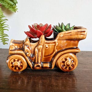 Op de afbeelding: Een bruine keramische vintage auto planter met twee vetplanten. De planter is gevormd als een klassieke auto met gedetailleerde wielen en carrosserie. Eén vetplant is rood, de andere groen.