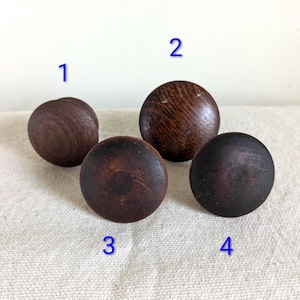 May include: Four wooden knobs in varying shades of brown are arranged on a light-colored surface. The knobs are round with a flat base, showcasing the natural wood grain and texture. These knobs could be used for furniture.