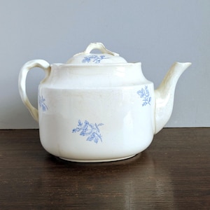 May include: Cream-colored ceramic teapot with a lid and spout. The teapot features blue floral designs and a curved handle. The teapot is sitting on a dark wooden surface.