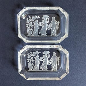 May include: Two clear, rectangular glass dishes with beveled edges. Each dish features an etched design of three figures and foliage. The figures appear to be in classical attire, carrying items. The dishes are displayed on a dark blue surface.