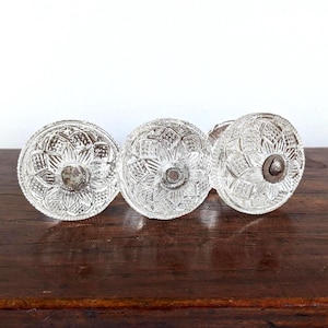 May include: Three circular glass cabinet knobs featuring a pressed geometric starburst pattern. Each knob has a clear, faceted surface and a central metal screw attachment point. The knobs are arranged in a horizontal row, showing their front faces.