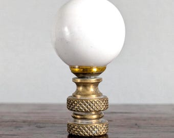 May include: A lamp finial featuring a smooth, spherical white orb mounted atop a metallic gold-toned base. The base consists of a small, polished gold collar connecting to a cylindrical, textured brass-colored stem with two knurled bands.