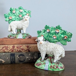 Op de afbeelding: Twee witte keramische schapenfiguren met groene en bruine details. Elk schaap staat onder een groene boom met rode en witte bloemen. De schapen staan op een groene en bruine basis.