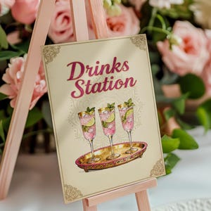 May include: A Drinks Station sign on a pink easel. The sign features the text "Drinks Station" in pink script, with illustrations of three cocktails on a gold tray. The cocktails have pink liquid, lime slices, and mint sprigs.