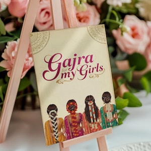Op de afbeelding: Decoratief bord op een kleine ezel met de tekst "Gajra for my Girls". Het bord toont illustraties van vier vrouwen met uitgebreide kapsels en kleurrijke traditionele kleding. De achtergrond bevat roze bloemen.