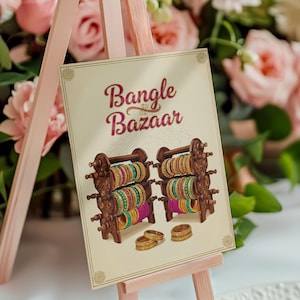 Op de afbeelding: Een bord op een roze ezel toont "Bangle Bazaar" in bordeauxrood schrift. Het bord toont illustraties van houten armbanddisplays met kleurrijke armbanden en gouden ringen. De achtergrond bevat roze bloemen.