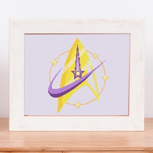 May include: A framed cross-stitch design featuring a yellow triangle with a purple swoosh and a tall, purple structure. The design is encircled by a yellow ring and stars. The frame is white, and the artwork is on a wooden surface.