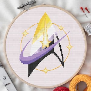 May include: A cross-stitch embroidery in a wooden hoop features a non-binary pride flag design. The design includes yellow, white, purple, and black elements, with a star and a circular border. Sewing supplies, including scissors, needles, and thread, are visible.