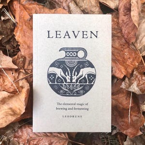 May include: A rectangular book with a light tan cover featuring the title LEAVEN in black text. Below the title is a black graphic of a ceramic vessel with two ibexes facing each other. The subtitle reads The elemental magic of brewing and fermenting.