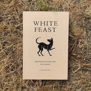 May include: A book titled "WHITE FEAST" with a black dog illustration on the cover. The book has a tan cover and is lying on a bed of dry grass. The text "Experimental summer-tide lore & praxis" and the publisher's name are also visible.