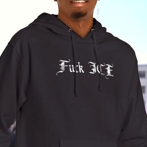 May include: A dark charcoal gray hooded sweatshirt featuring a front kangaroo pocket and long sleeves. The chest displays the text "Fuck ICE" printed in a white, stylized gothic font. The garment includes a matching drawstring hood.