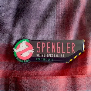May include: A rectangular Spengler name badge with a Ghostbusters logo and the text "SLIME SPECIALIST NEW YORK UNIT". The badge is black with pink and yellow accents and is pinned to a red and black plaid shirt.